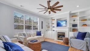 A spacious living room with a fireplace, a ceiling fan, and a large window. - Coastal Quarters in Bluffton - Dog Friendly! Bluffton Park Lowcountry Charmer (Bluffton)