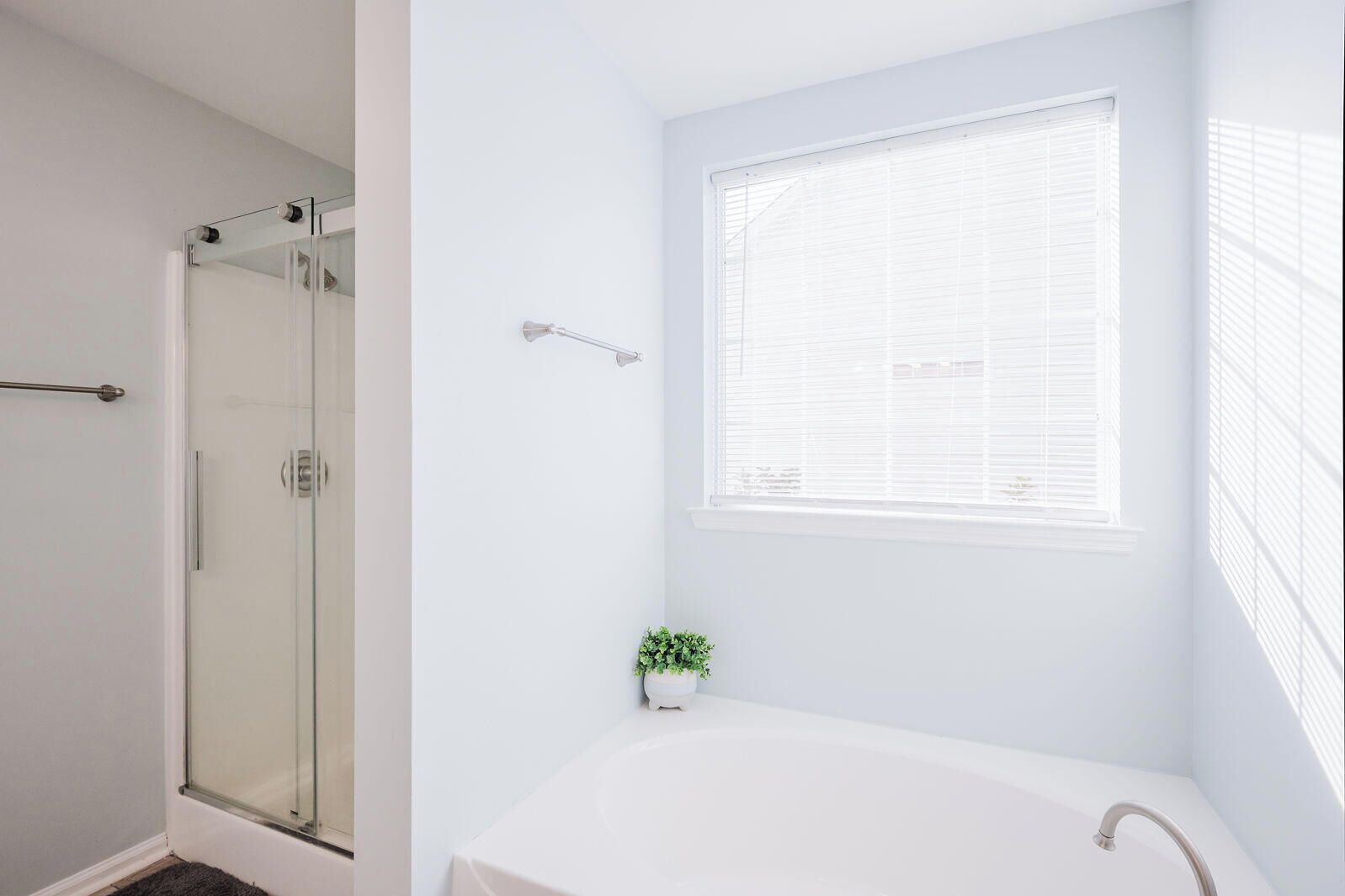 A modern bathroom with a glass shower enclosure, a white bathtub, and a window with blinds.