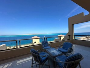 A balcony with wicker chairs and a glass table overlooking a beach and ocean.