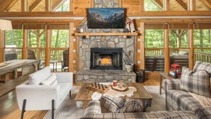 A cozy cabin interior with a stone fireplace, wooden furniture, and large windows offering a view of the forest. - DreamForest 3BR, Firepit, Foosball, Banner Elk (Banner Elk)