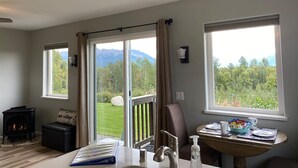 A room with a fireplace, a dining table with chairs, a view of a green landscape through large windows, and a sliding glass door leading to an outdoor area.