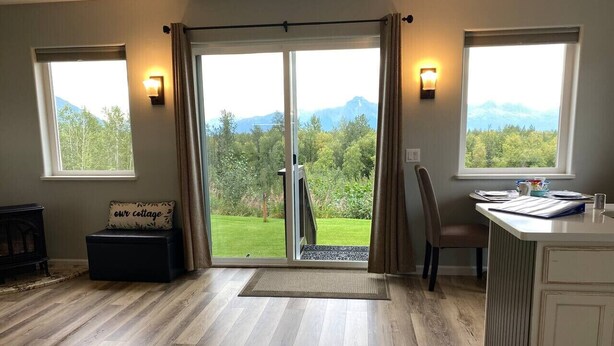A room with a large sliding glass door leading to a balcony, a wooden floor, a small seating area with a cushion, and a kitchenette area with a counter and cabinets.