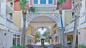 The entrance to The Village of Baytowne Wharf with a central archway, palm trees, and a brick pathway. - Tivoli by the Sea 5286 (Miramar Beach)
