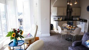 A modern living room with a sofa, a dining area with a table and chairs, and a kitchen with cabinets and appliances.
