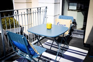 Une table bleue placée à l’extérieur, sur laquelle se trouve un objet jaune, deux chaises bleues avec des coussins, et un tapis à rayures sur un balcon.