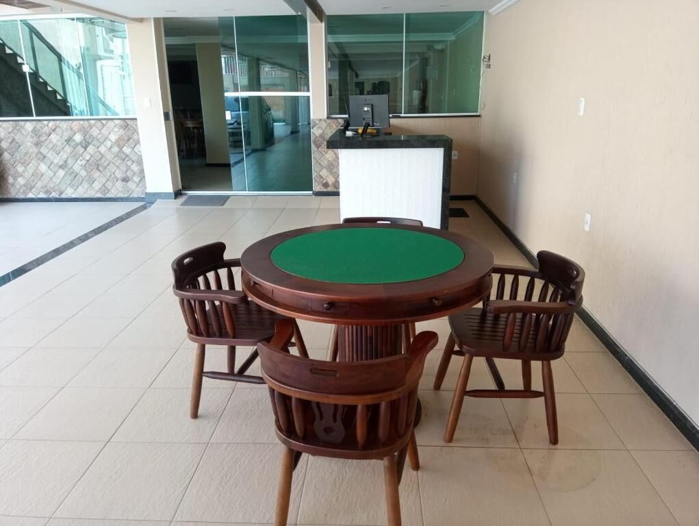 Una mesa redonda de madera con superficie de fieltro verde y cuatro sillas de madera en un área de recepción con suelo de baldosas y una pared de vidrio al fondo.