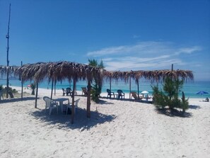 Une plage avec des parasols en chaume, des chaises en plastique blanches et un ciel bleu azur.