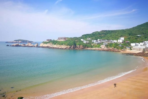 A coastal beach with a sandy shore, a clear sky, and buildings on a hillside.