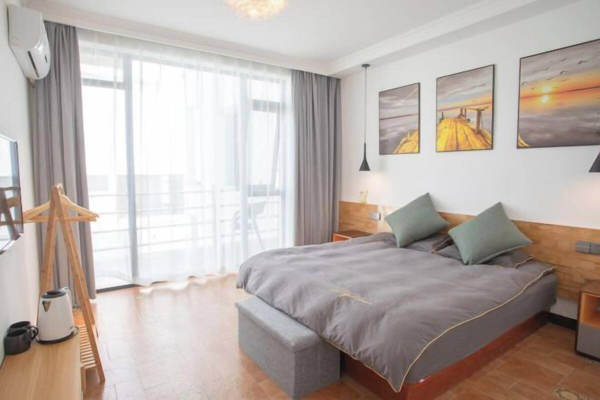 A hotel room with a large bed, a wooden headboard, a bench, a coffee maker, and a view of the outdoors through large windows.