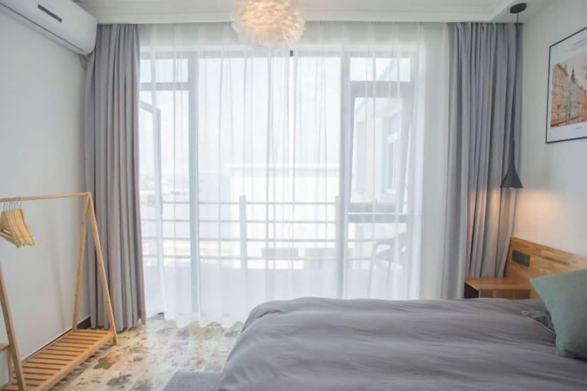 A bedroom with a bed, a wooden headboard, a hanging light fixture, a window with sheer curtains, and a wooden floor.