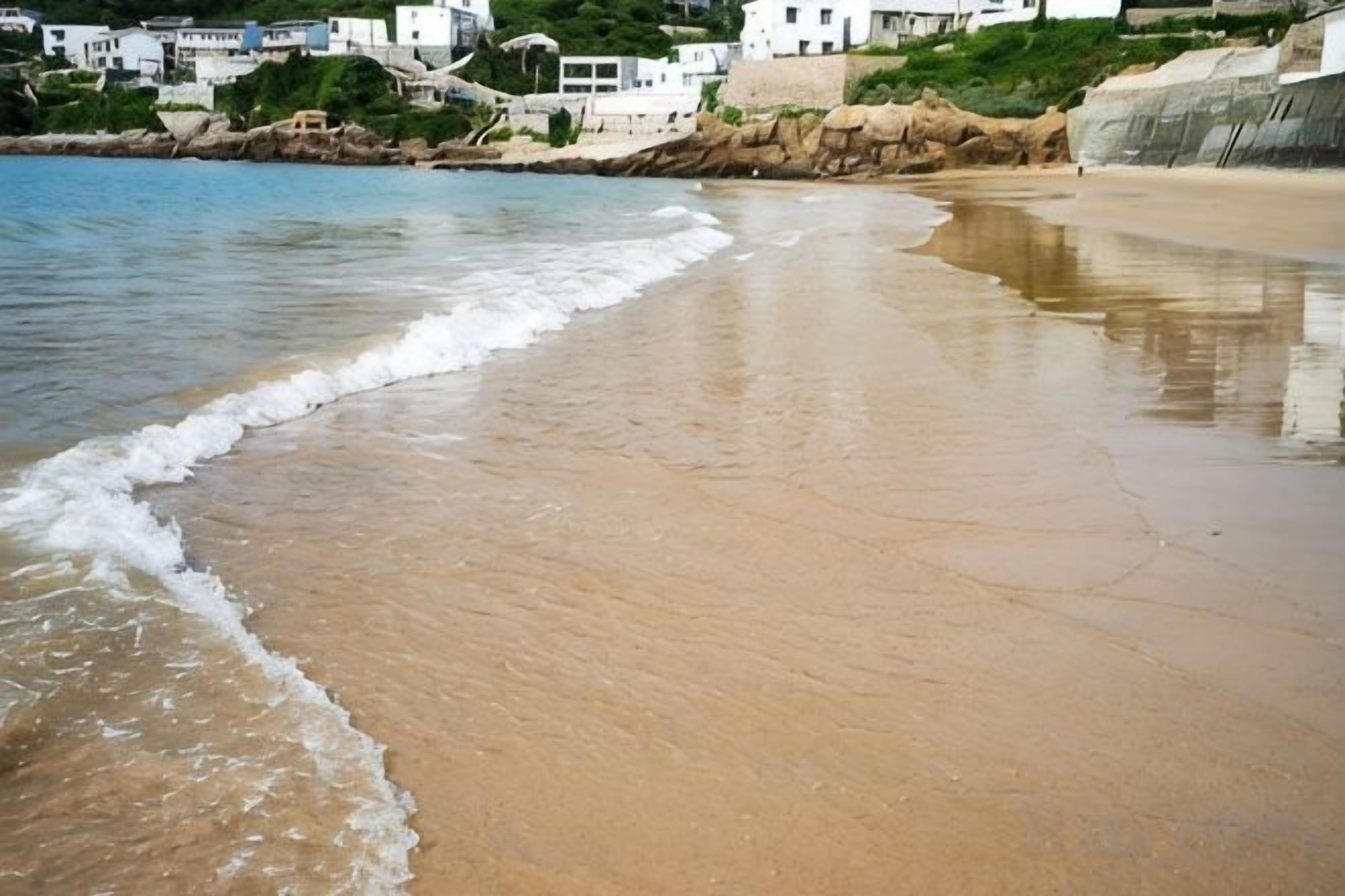 A beach with waves, a sandy shore, and buildings in the background.