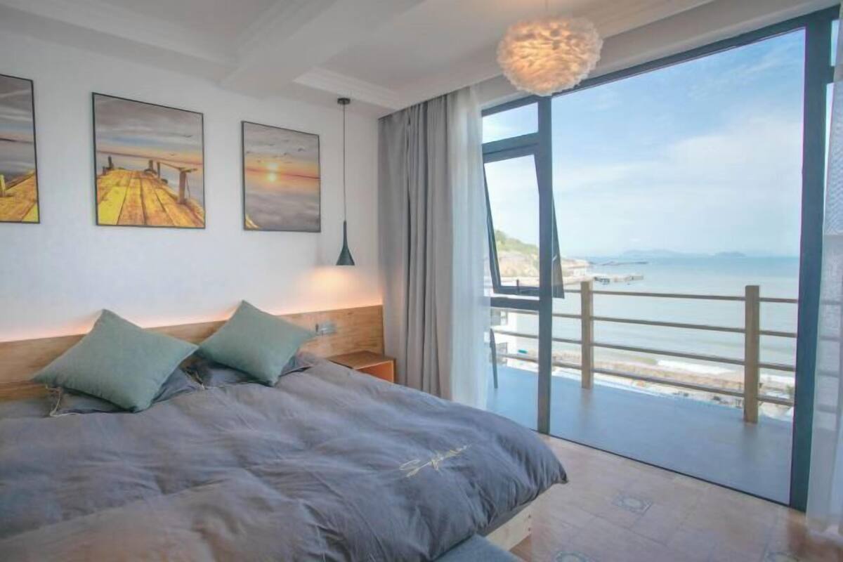 A bedroom with a large bed, a view of the sea through a balcony, and artwork on the wall.