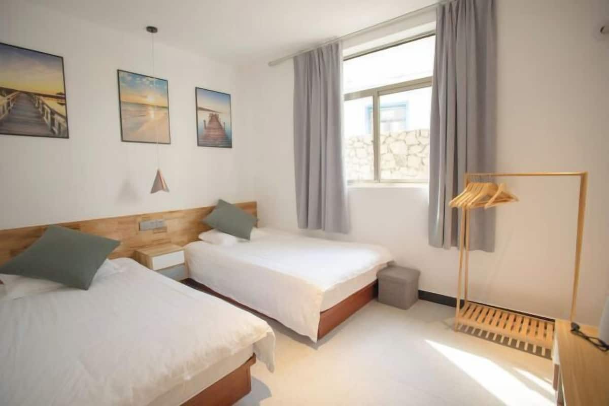 A hotel room with two beds, a window with curtains, a wooden floor, and a wall-mounted lamp.