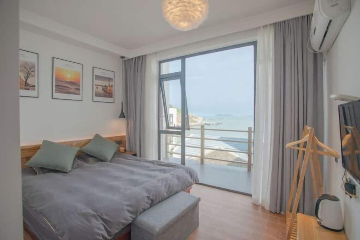 A hotel room with a bed, a bench, a TV, and a view of the sea.