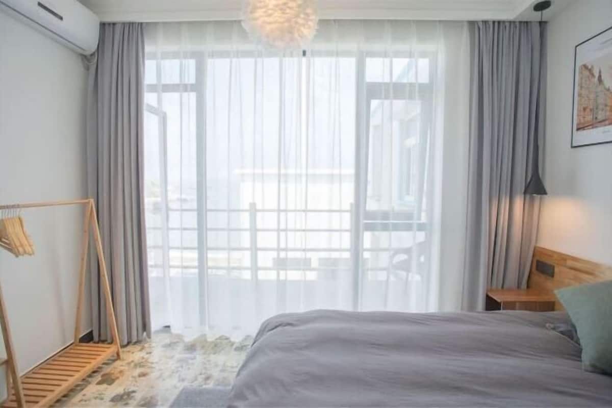 A bedroom with a bed, a wooden headboard, a hanging light fixture, a wooden bench, and a view through a window with curtains.