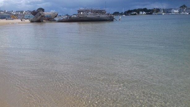 Ein Strand mit klarem, flachem Wasser, einem großen Boot und einem Steg mit Gebäuden im Hintergrund.