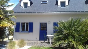 Ein zweigeschossiges Haus mit dunklem Dach, weißen Wänden und blauen Fensterläden. Im zweiten Stock befindet sich ein kleines Fenster und ein mit einem blauen Fensterladen versehenes Haus. Vor dem Haus liegt eine kleine Terrasse mit Tisch und Stühlen.