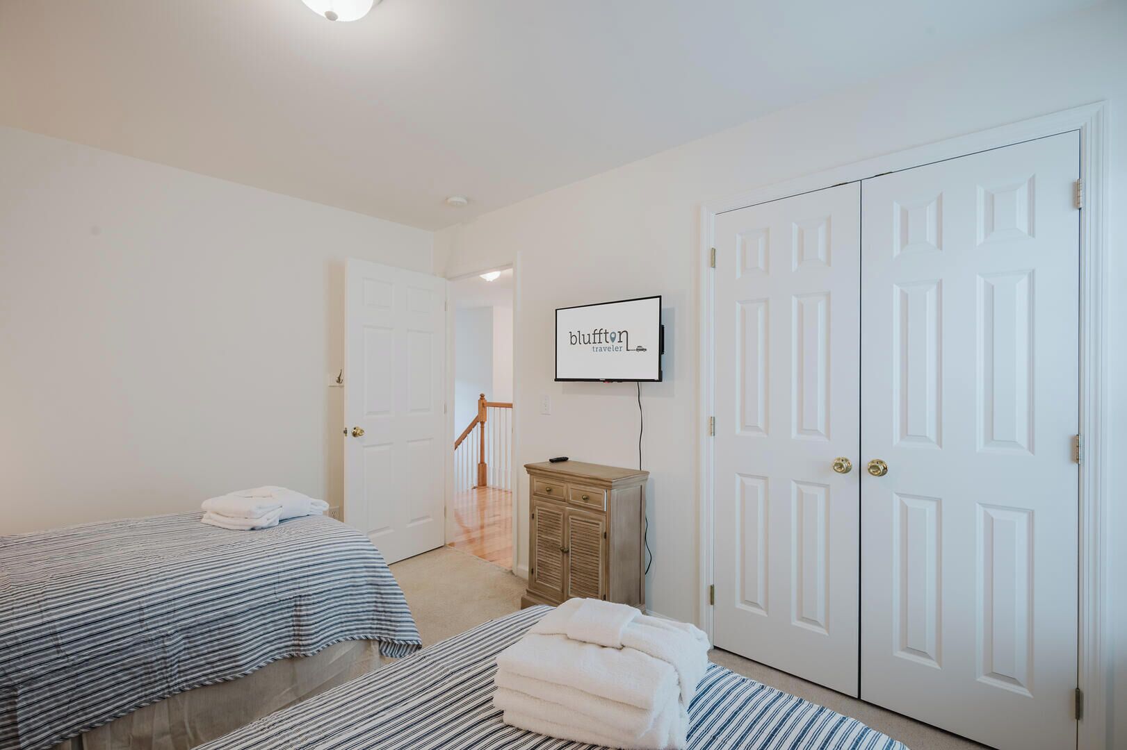 A hotel room with two beds, a wooden dresser, a television, and a door with a 'bluffton traveler' sign.