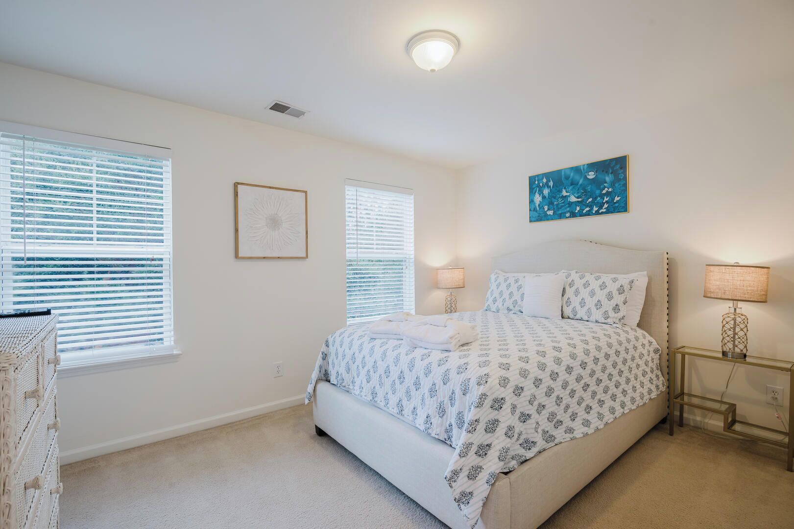 A bedroom with a bed, two bedside tables with lamps, a wall art piece, and two windows with blinds.