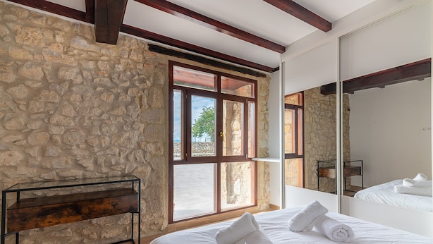 A bedroom with a stone wall, wooden beams, a large window, a bed with white linens, and a bench. - Infinity Coast I by Fidalsa (Miengo)