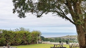 A picnic area with tables and benches, overlooking a coastal landscape with a view of the sea and a distant town. - Infinity Coast I by Fidalsa (Miengo)