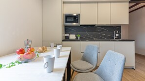 Une cuisine moderne avec des armoires blanches, un comptoir en marbre noir et un four à micro-ondes encastré. On y trouve deux chaises grises et une table sur laquelle se trouve un saladier rempli de fruits et plusieurs tasses blanches.