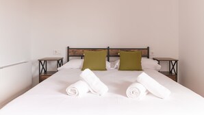 Un lit bien fait, avec du linge de lit blanc et deux serviettes enroulées, une tête de lit en bois et deux tables de chevet ornées de coussins verts.