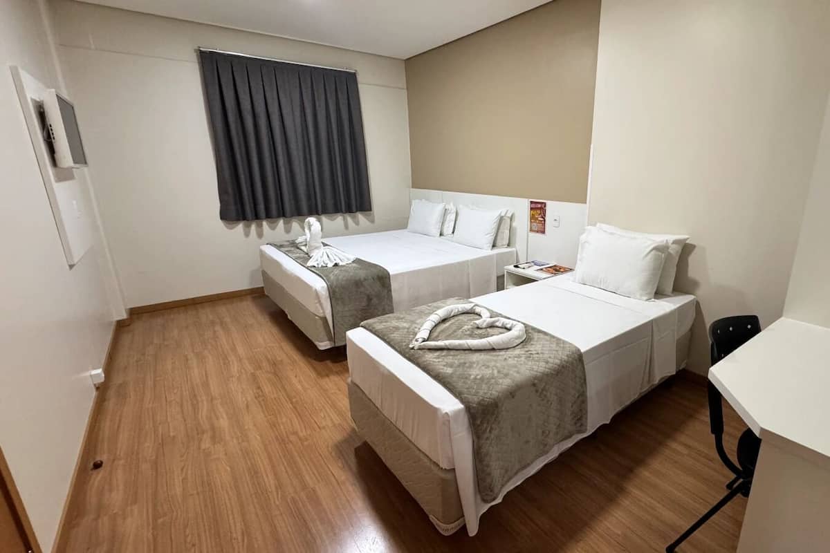 Une chambre d’hôtel avec deux lits simples, une petite table de chevet avec une lampe, un bureau avec une chaise et une fenêtre avec des rideaux.
