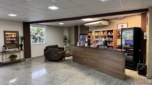 A reception area with a counter, seating, a large window, and a vending machine. - Hotel Zandona (Blumenau)