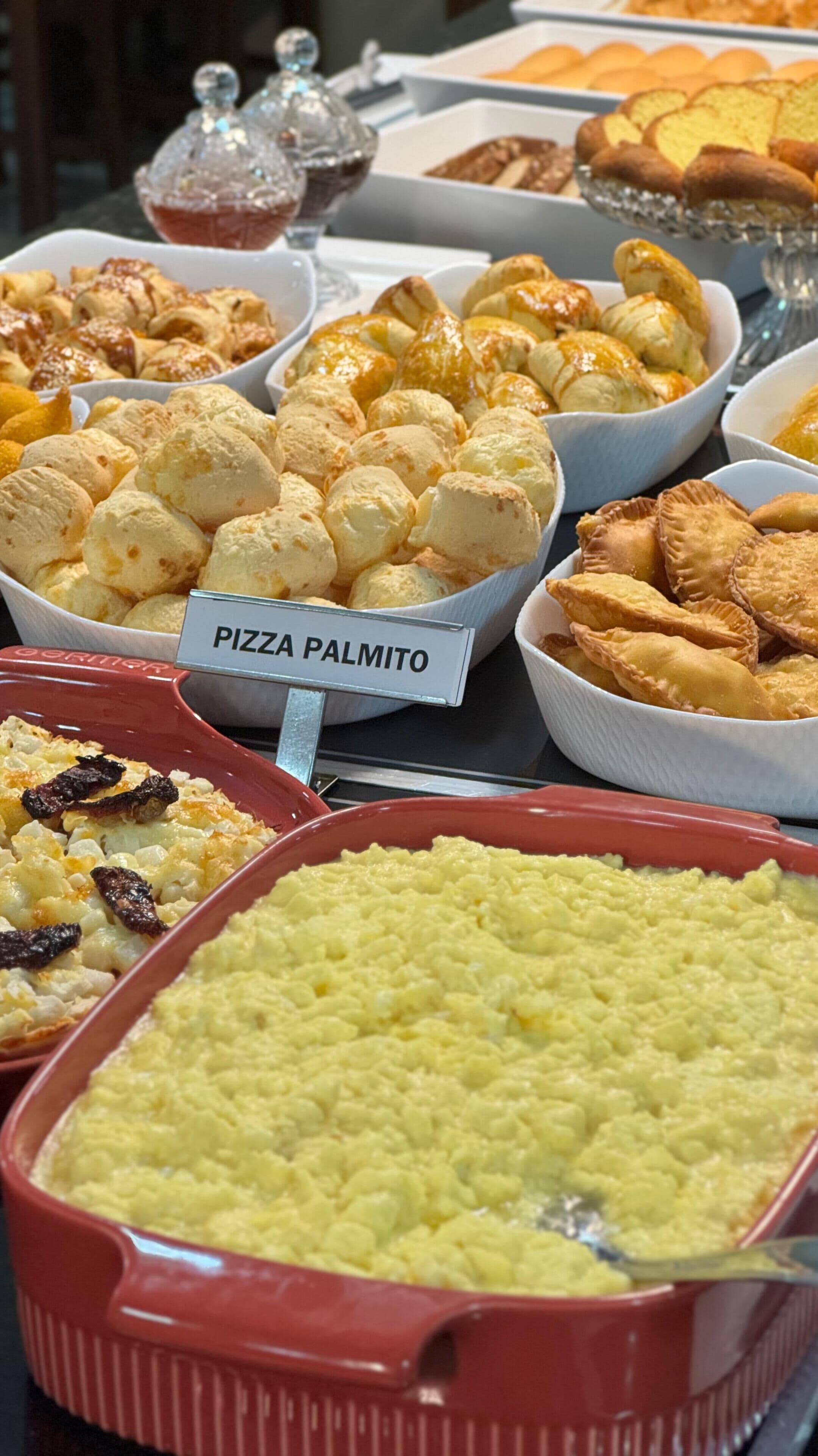 Un bufé con diversas opciones de comida, incluyendo pizza de palmito, pastelería y un plato al horno.