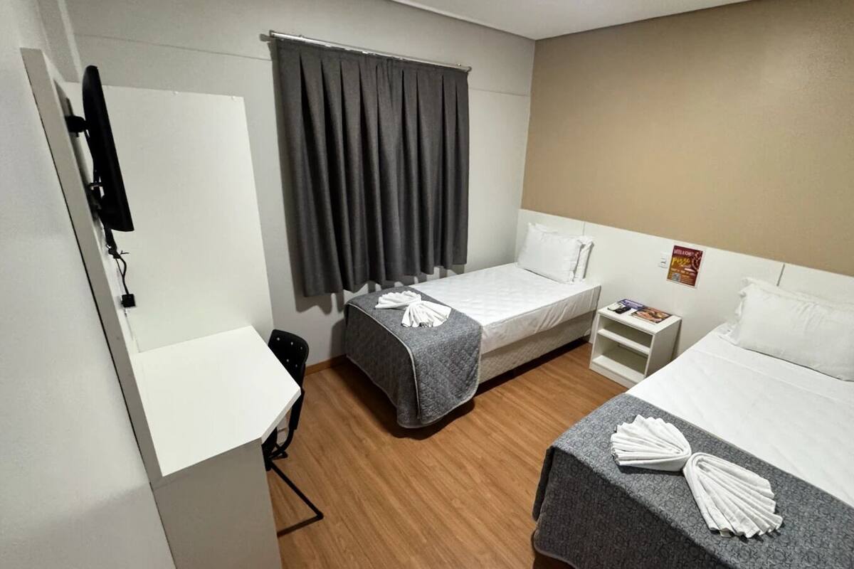 Une chambre d’hôtel avec deux lits simples, un bureau, une chaise, une petite étagère avec un magazine et un téléviseur fixé au mur.