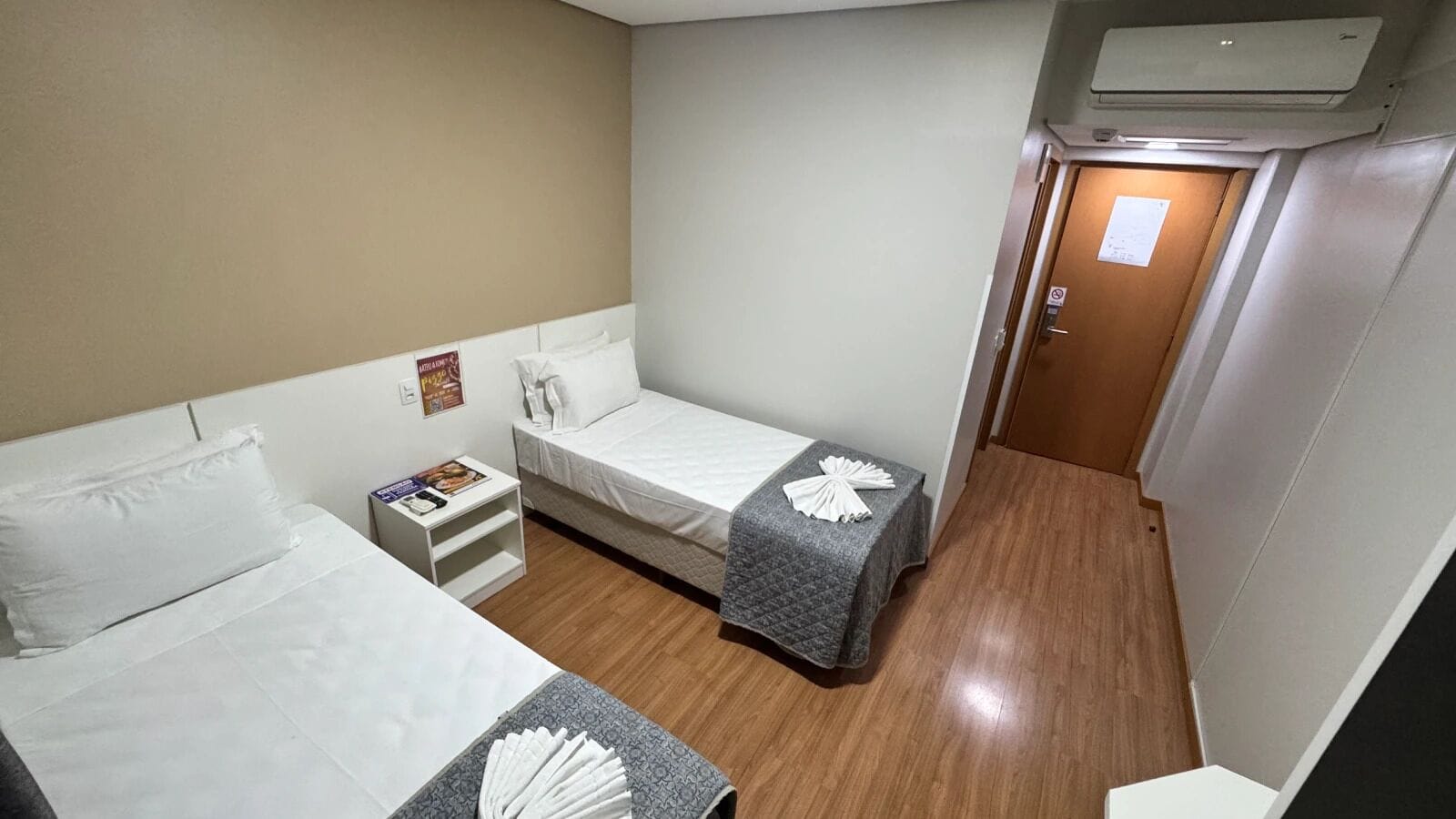 Quarto pequeno e limpo, com duas camas, uma mesinha de cabeceira, uma pequena mesa e um ar condicionado.