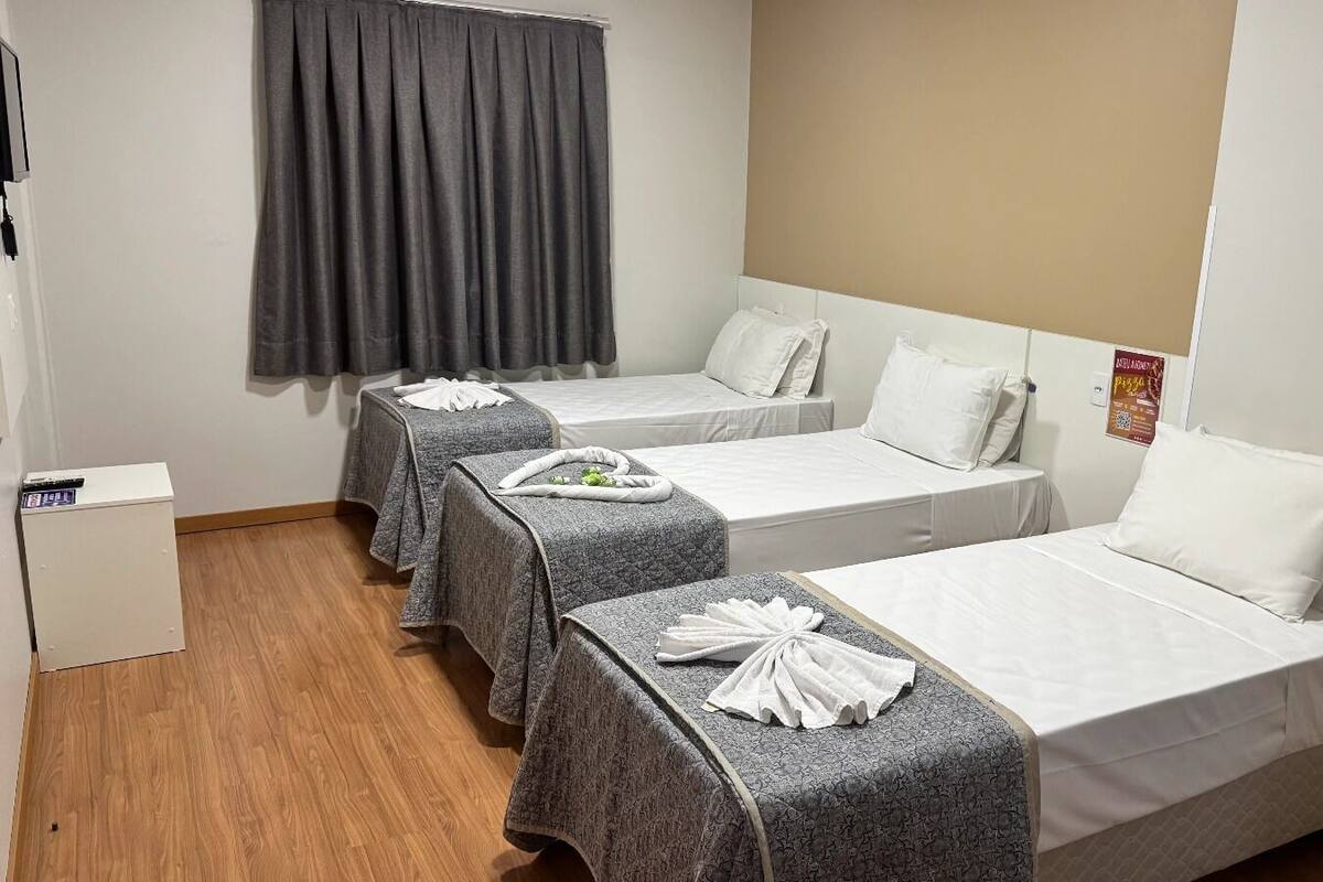 Une chambre d’hôtel avec trois lits simples, chacun recouvert d’une couverture grise et agrémenté d’oreillers blancs. La chambre contient également une petite armoire blanche et une chaise noire.