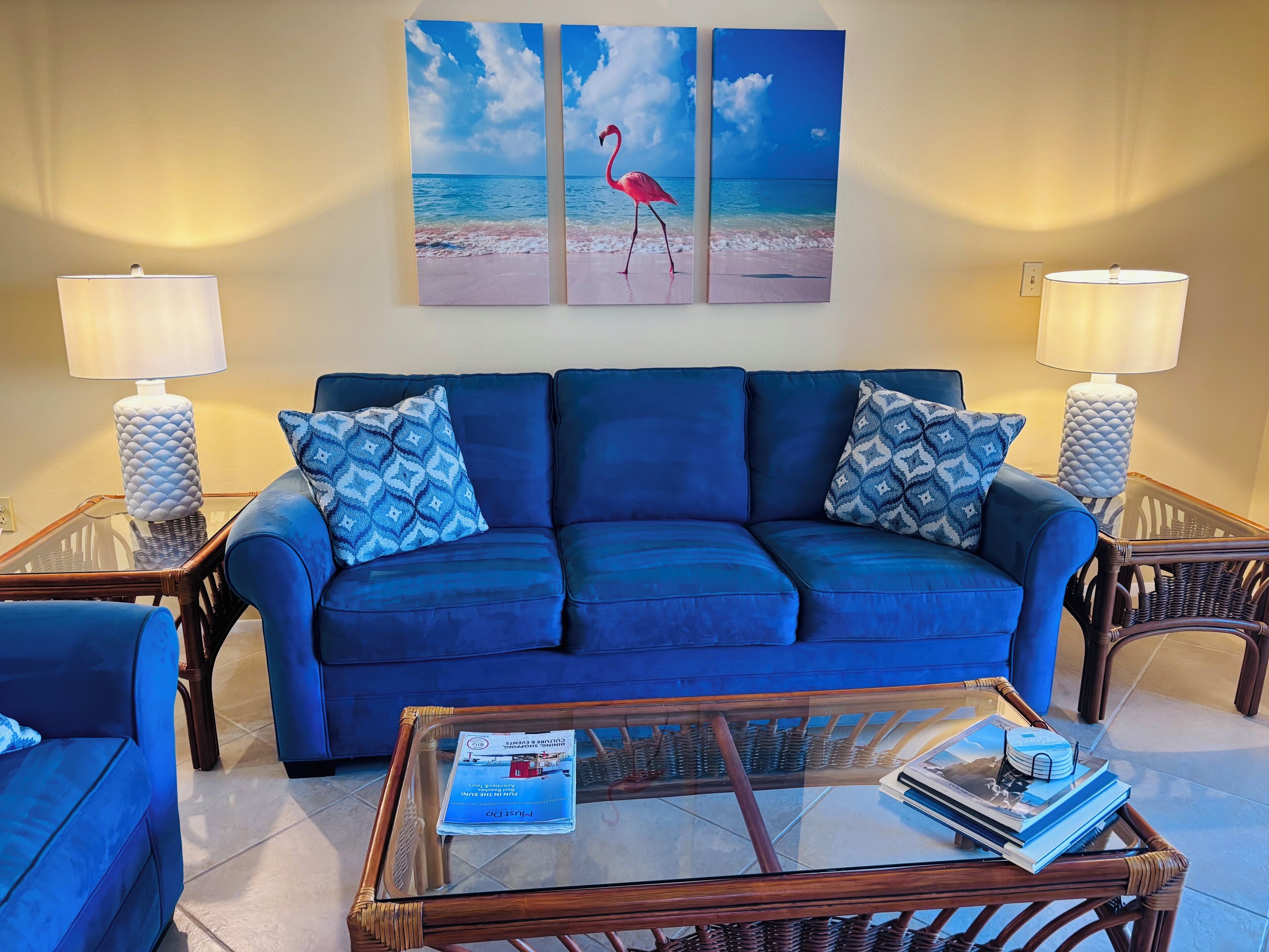 Una sala de estar con un sofá azul, almohadones estampados, una mesa de centro de vidrio con revistas y una obra de arte en la pared que representa una escena de playa con un flamenco.