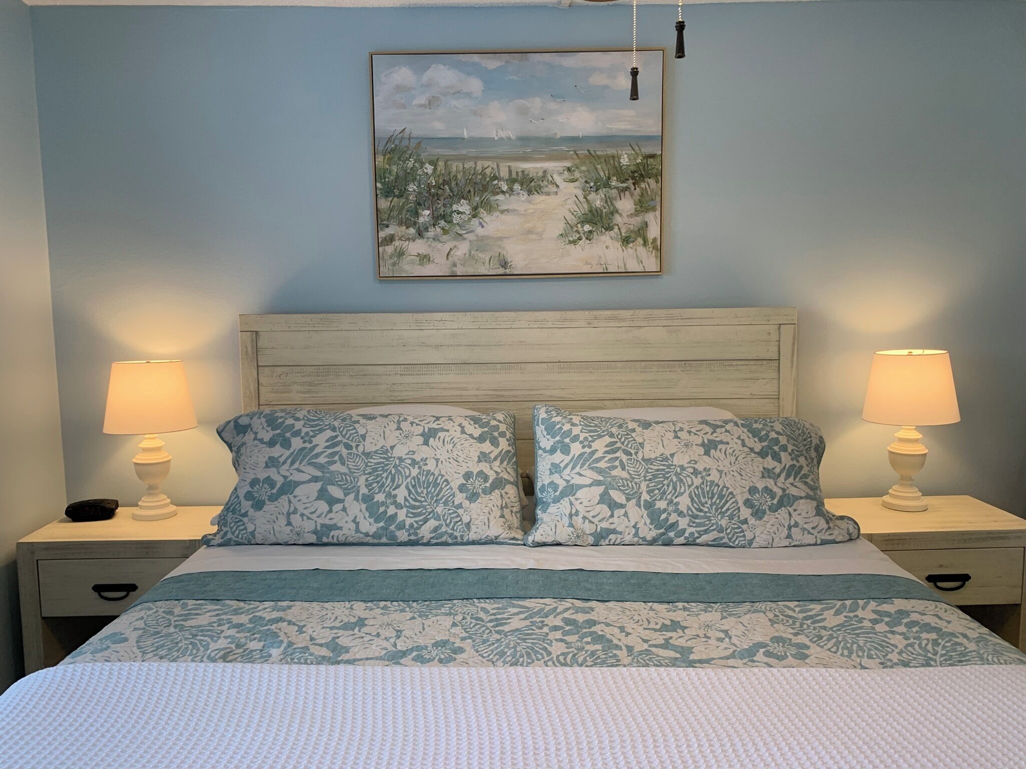 Une chambre à coucher avec un lit, deux lampes de chevet, une table de chevet, un tiroir et un tableau au mur.