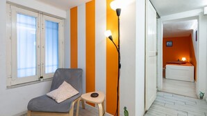 Une pièce avec une chaise grise, un petit tabouret, une lampe sur pied et un lit à l’arrière-plan.