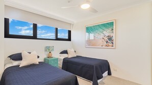 Un dormitorio con dos camas, un ventilador de techo, una ventana con vista al cielo y un cuadro en la pared.