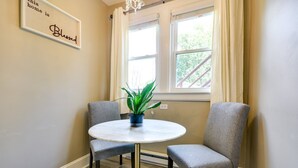 A dining area with a round table, two grey upholstered chairs, a potted plant, and a window with curtains.