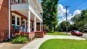 A two-story brick house with a covered porch, a lawn, and a red car parked on the street.