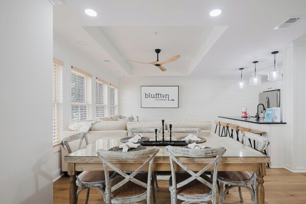 A spacious living room with a large white sofa, a wooden dining table with four chairs, and a ceiling fan.
