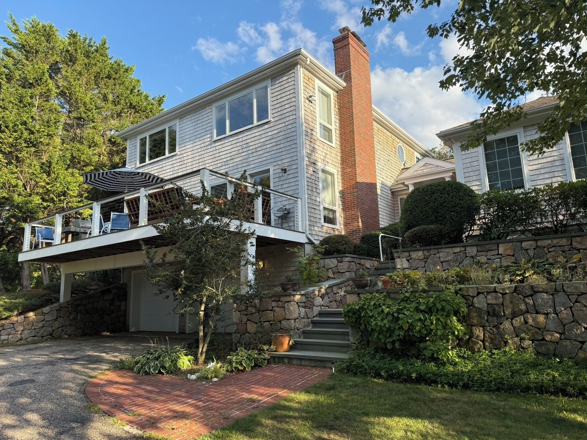A two-story house with a stone retaining wall, a covered deck with chairs, and a brick pathway leading to the garage.