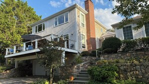 A two-story house with a stone retaining wall, a covered deck with chairs, and a brick pathway leading to the garage.
