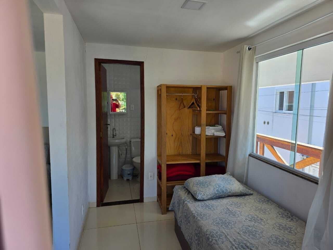 Une petite pièce bien éclairée, avec un lit, une armoire en bois et une fenêtre donnant sur des immeubles.