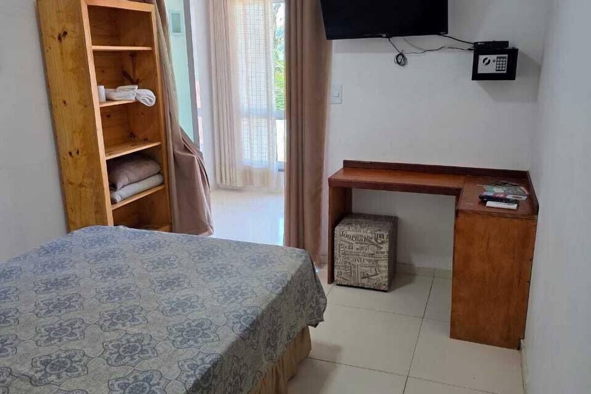 A hotel room with a bed, a wooden wardrobe, a desk with a chair, a television, and an air conditioning unit.