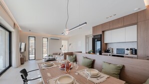Un comedor moderno con una mesa de madera preparada para cuatro personas, una cocina al fondo y vistas al exterior a través de amplios ventanales.