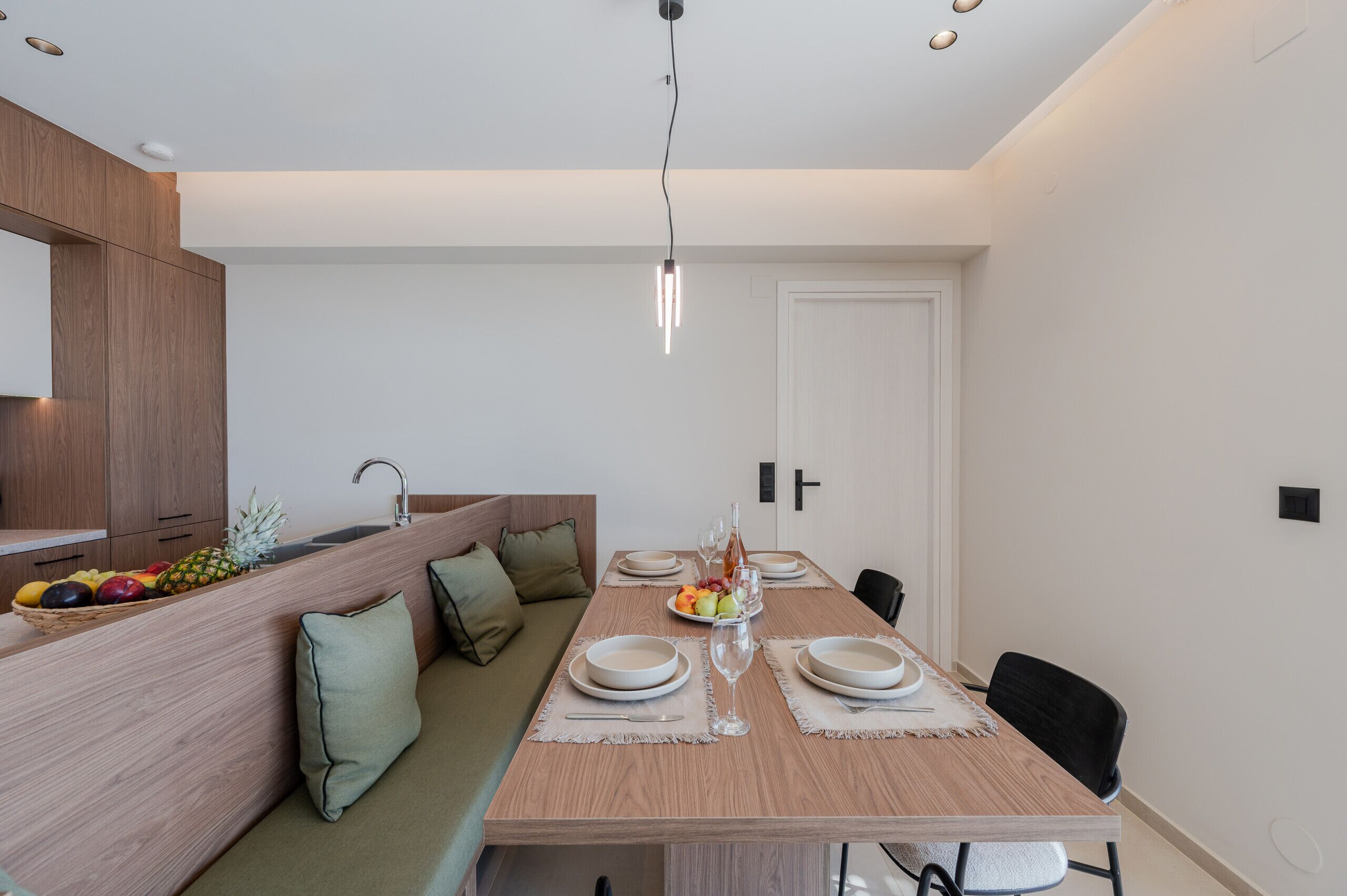 Un espace repas moderne avec une table en bois dressée pour quatre personnes, un coin cuisine agrémenté d’un panier de fruits et un banc intégré avec des coussins.