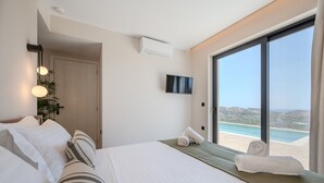 A hotel room with a bed, a TV, a large window with a beach view, and a plant. - Filade Luxury Villa 2 (Agios Vasileios)