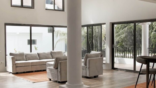 A spacious living area with a large sofa set, a coffee table, and a dining table. There are large windows providing natural light and views of palm trees. - The Escape Ghana - Garden Villa (Accra)