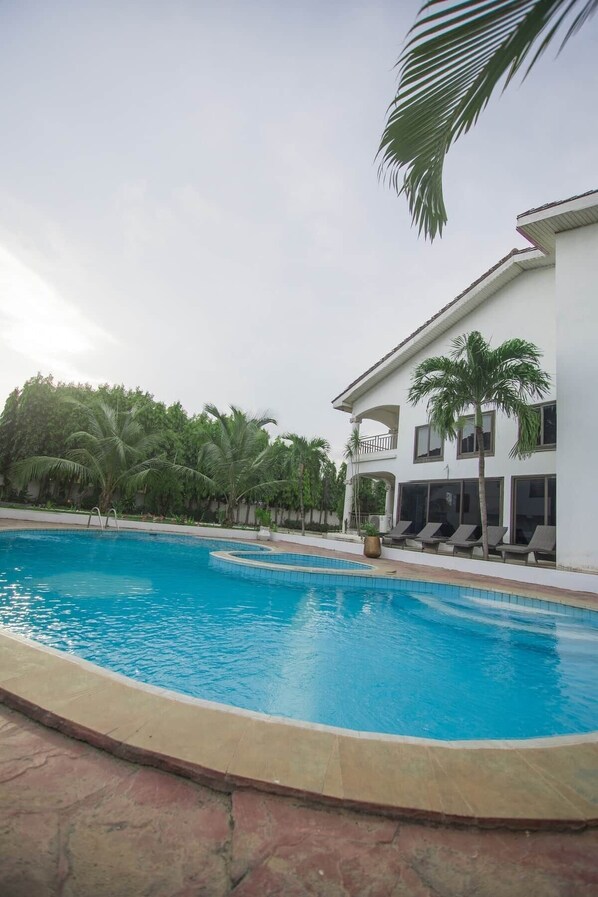 A swimming pool with a curved edge, surrounded by palm trees and a white building with a balcony.