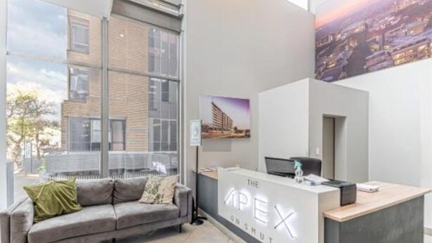A reception area with a modern design, featuring a reception desk labeled "THE APEX ONSHUTTS" and a grey sofa with cushions.
